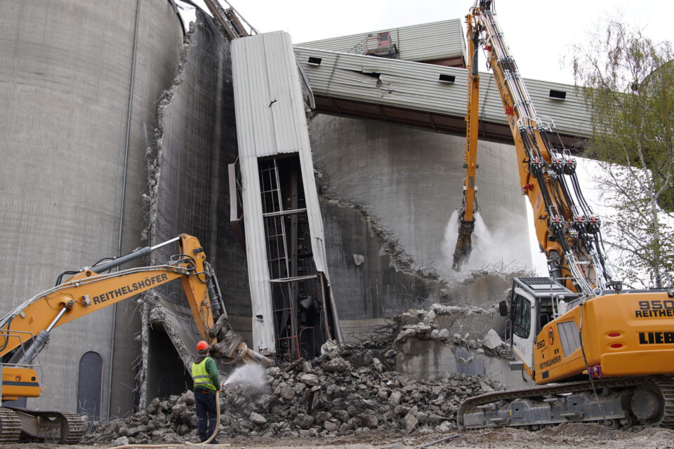 Das Bild zeigt den Abriss eines großen Betonbauwerks mit zwei schweren Baggern, die Trümmer abtragen. Ein Arbeiter in Schutzkleidung steht vor dem Schutt und hält einen Schlauch, während Staub aufwirbelt. Die eingestürzten Betonwände und freiliegenden Stahlstreben verdeutlichen den fortgeschrittenen Zustand der Abbrucharbeiten.