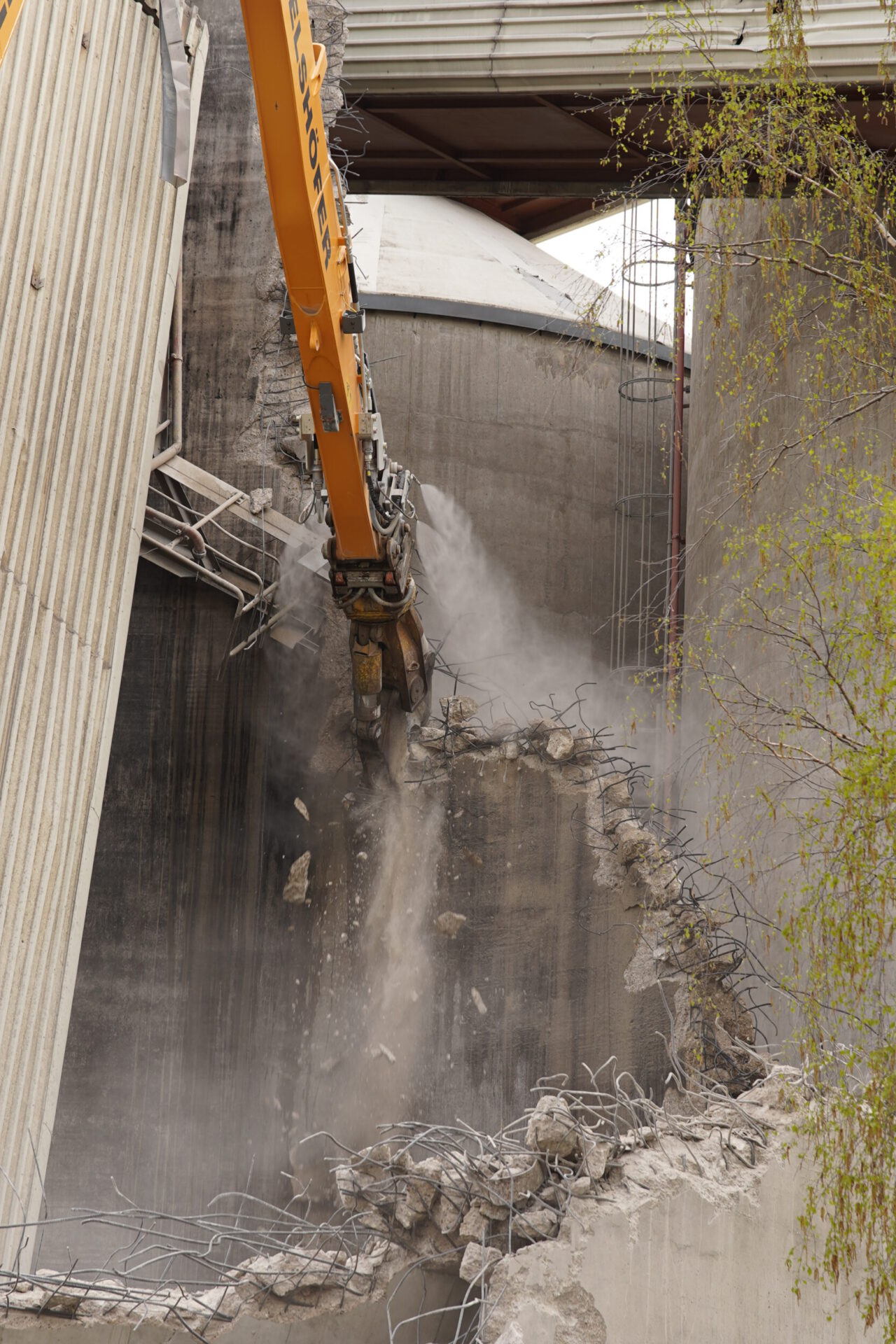 Das Bild zeigt den Greifarm eines großen Baggers, der ein Loch in eine massive Betonwand bricht. Betonstücke fallen herab, während Staub und freiliegende Stahlbewehrungen sichtbar werden. Im Hintergrund sind weitere Teile der Industrieanlage und ein Baum zu erkennen.