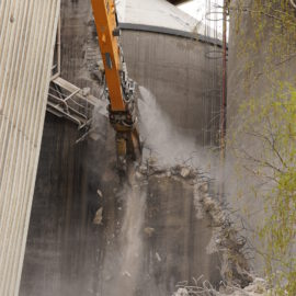 Das Bild zeigt den Greifarm eines großen Baggers, der ein Loch in eine massive Betonwand bricht. Betonstücke fallen herab, während Staub und freiliegende Stahlbewehrungen sichtbar werden. Im Hintergrund sind weitere Teile der Industrieanlage und ein Baum zu erkennen.