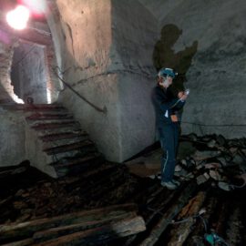 Das Bild zeigt eine Person mit Schutzhelm, die in einem dunklen, unterirdischen Gewölberaum steht und mit einem Messgerät arbeitet. Der Boden ist teilweise eingestürzt und mit Schutt sowie Holzbalken bedeckt. Eine Treppe und gemauerte Wände deuten auf ein älteres Bauwerk hin, das untersucht wird.