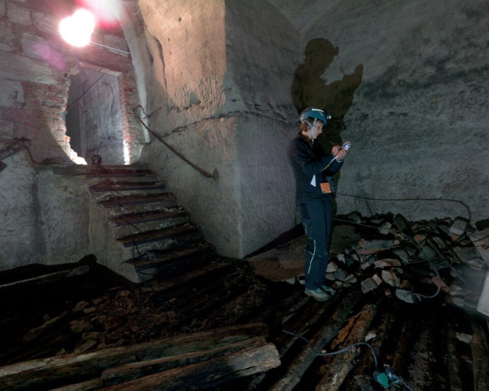 Das Bild zeigt eine Person mit Schutzhelm, die in einem dunklen, unterirdischen Gewölberaum steht und mit einem Messgerät arbeitet. Der Boden ist teilweise eingestürzt und mit Schutt sowie Holzbalken bedeckt. Eine Treppe und gemauerte Wände deuten auf ein älteres Bauwerk hin, das untersucht wird.