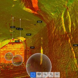 Das Bild zeigt eine farblich codierte 3D-Darstellung eines unterirdischen Hohlraums oder Tunnels mit deutlich erkennbaren Wand- und Deckenstrukturen. Mehrere markierte Punkte und Messlinien weisen auf Risse, Abstände und Auffälligkeiten hin. Die Darstellung wirkt wie eine technische Analyse zur Bauwerksprüfung oder Schadenserfassung.