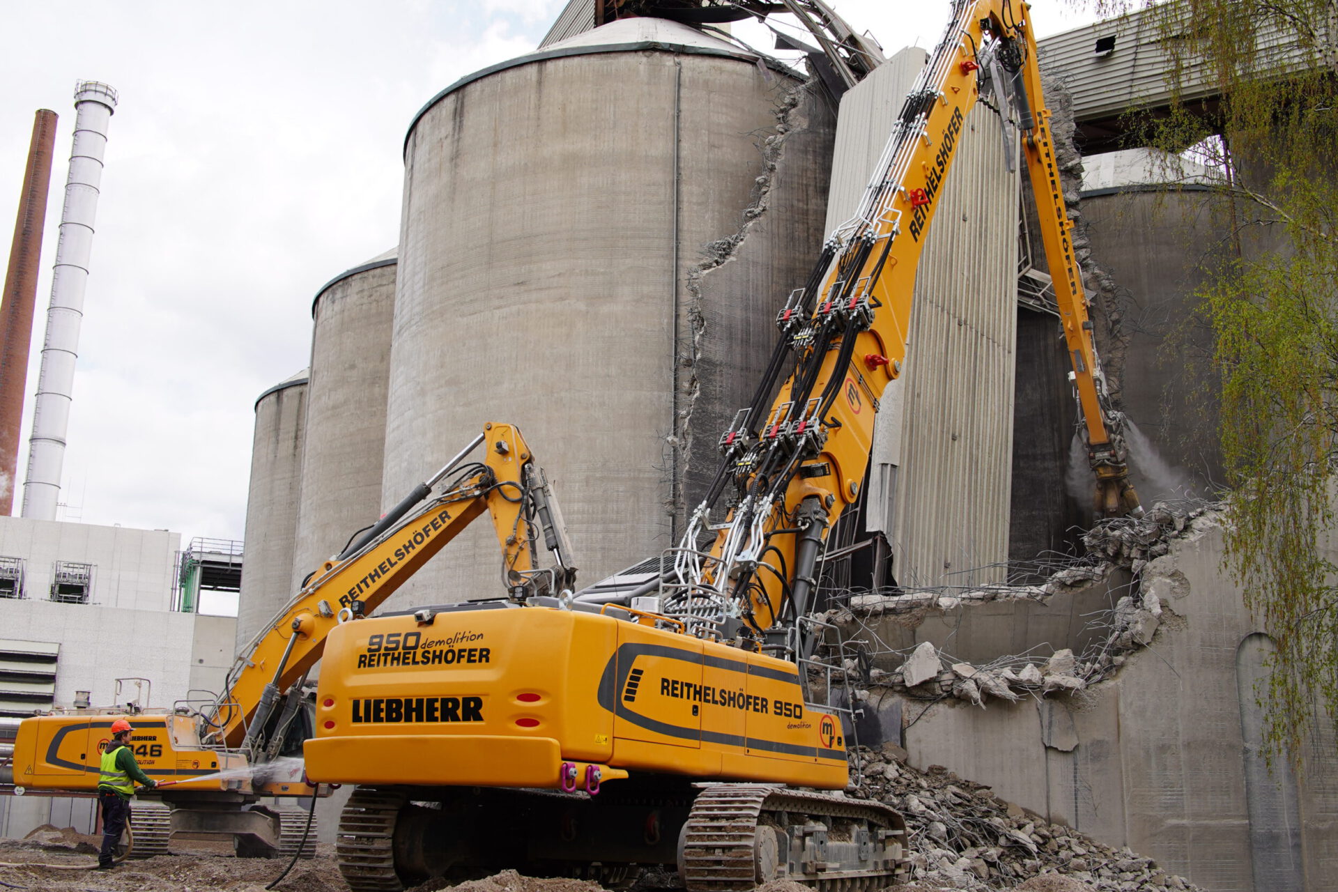 Das Bild zeigt zwei große Bagger, die an einem massiven Betonsilo Abbrucharbeiten durchführen. Betonbrocken und freiliegende Stahlstreben liegen als Schutt am Boden, während eine Person mit Schutzkleidung seitlich arbeitet. Im Hintergrund sind weitere Silos und industrielle Anlagen zu erkennen.