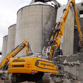 Das Bild zeigt zwei große Bagger, die an einem massiven Betonsilo Abbrucharbeiten durchführen. Betonbrocken und freiliegende Stahlstreben liegen als Schutt am Boden, während eine Person mit Schutzkleidung seitlich arbeitet. Im Hintergrund sind weitere Silos und industrielle Anlagen zu erkennen.