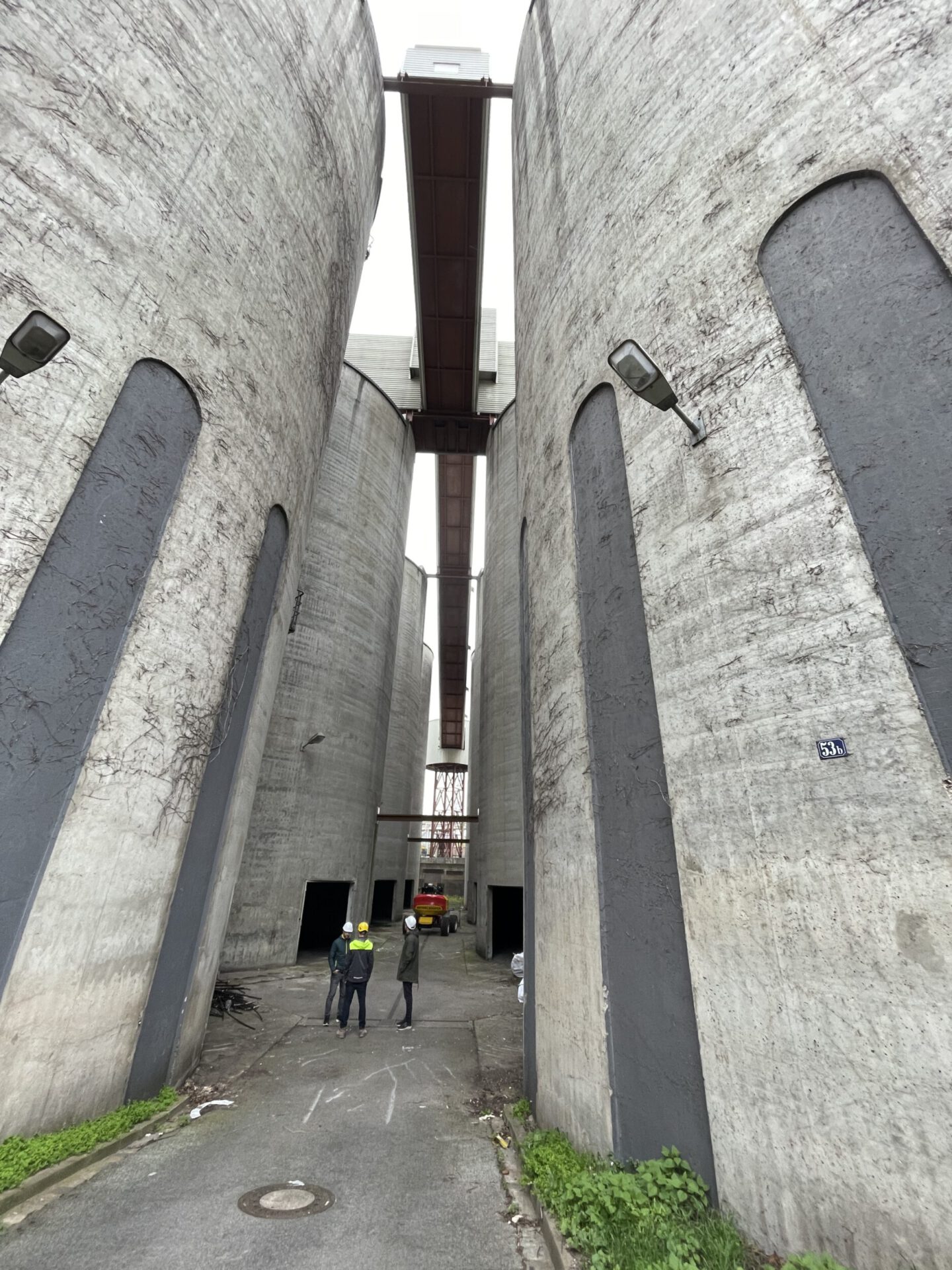 Das Bild zeigt einen schmalen Durchgang zwischen hohen, zylindrischen Betonsilos. Drei Personen mit Schutzhelmen stehen auf dem Boden und blicken in Richtung eines roten Fahrzeugs im Hintergrund. Über den Silos verlaufen mehrere Brücken und Metallkonstruktionen, die die Bauwerke miteinander verbinden.