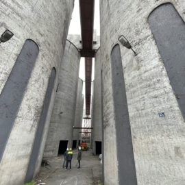 Das Bild zeigt einen schmalen Durchgang zwischen hohen, zylindrischen Betonsilos. Drei Personen mit Schutzhelmen stehen auf dem Boden und blicken in Richtung eines roten Fahrzeugs im Hintergrund. Über den Silos verlaufen mehrere Brücken und Metallkonstruktionen, die die Bauwerke miteinander verbinden.