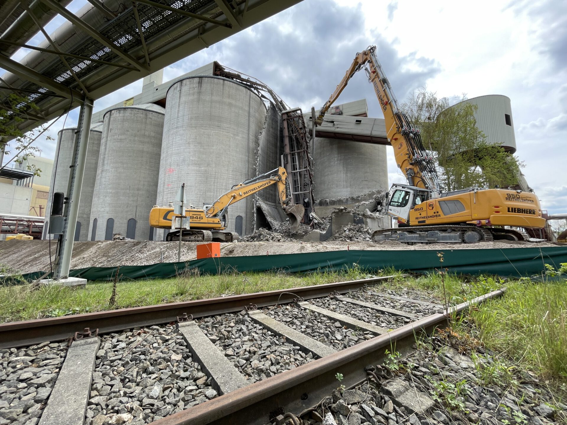 Das Bild zeigt zwei große Bagger, die an mehreren massiven Betonsilos Abbrucharbeiten durchführen. Im Vordergrund verlaufen Bahnschienen über Schotter, dahinter liegt ein abgesperrter Arbeitsbereich mit Schutt und Trümmern. Über der Baustelle sind Rohrleitungen und Metallkonstruktionen zu sehen.