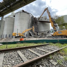 Das Bild zeigt zwei große Bagger, die an mehreren massiven Betonsilos Abbrucharbeiten durchführen. Im Vordergrund verlaufen Bahnschienen über Schotter, dahinter liegt ein abgesperrter Arbeitsbereich mit Schutt und Trümmern. Über der Baustelle sind Rohrleitungen und Metallkonstruktionen zu sehen.