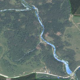 Das Bild zeigt eine Luftaufnahme einer Landschaft mit einem schmalen Fluss, der sich in einem geschwungenen Verlauf durch das Gebiet zieht. Der Fluss ist in einem kräftigen Blauton hervorgehoben, während die umliegenden Flächen überwiegend grün sind und Wiesen, Felder sowie bewaldete Bereiche erkennen lassen. Entlang des Flusses verlaufen Straßen, und es sind vereinzelte Strukturen sichtbar, die auf eine ländliche Umgebung mit Infrastruktur hinweisen.