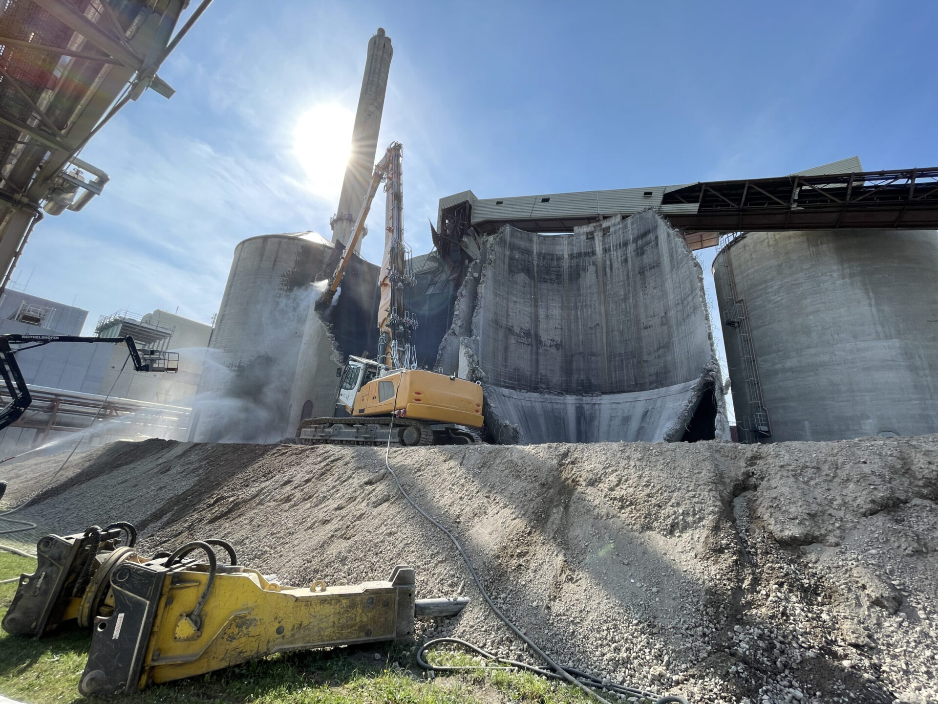 Das Bild zeigt den Abbruch eines großen zylindrischen Silos aus Beton, das von einem schweren Bagger mit langem Abbrucharm bearbeitet wird. Staub steigt aus der aufgebrochenen Wand auf, während Schutt auf einem hohen Erdhügel liegt. Im Vordergrund sind weitere Abbruchwerkzeuge und Kabel auf dem Boden zu erkennen.