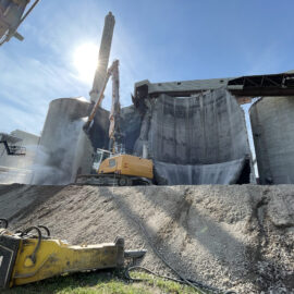 Das Bild zeigt den Abbruch eines großen zylindrischen Silos aus Beton, das von einem schweren Bagger mit langem Abbrucharm bearbeitet wird. Staub steigt aus der aufgebrochenen Wand auf, während Schutt auf einem hohen Erdhügel liegt. Im Vordergrund sind weitere Abbruchwerkzeuge und Kabel auf dem Boden zu erkennen.