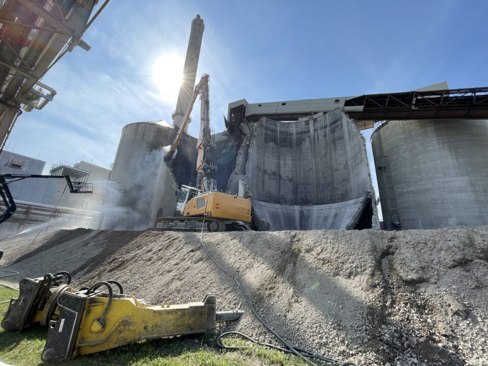 Das Bild zeigt den Abbruch eines großen zylindrischen Silos aus Beton, das von einem schweren Bagger mit langem Abbrucharm bearbeitet wird. Staub steigt aus der aufgebrochenen Wand auf, während Schutt auf einem hohen Erdhügel liegt. Im Vordergrund sind weitere Abbruchwerkzeuge und Kabel auf dem Boden zu erkennen.