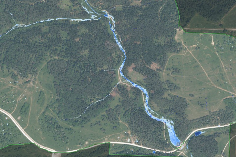 Das Bild zeigt eine Luftaufnahme einer Landschaft mit einem schmalen Fluss, der sich in einem geschwungenen Verlauf durch das Gebiet zieht. Der Fluss ist in einem kräftigen Blauton hervorgehoben, während die umliegenden Flächen überwiegend grün sind und Wiesen, Felder sowie bewaldete Bereiche erkennen lassen. Entlang des Flusses verlaufen Straßen, und es sind vereinzelte Strukturen sichtbar, die auf eine ländliche Umgebung mit Infrastruktur hinweisen.