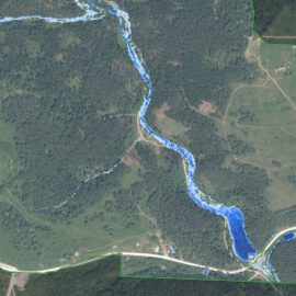 Das Bild zeigt eine Luftaufnahme einer Landschaft mit einem schmalen Fluss, der sich in einem geschwungenen Verlauf durch das Gebiet zieht. Der Fluss ist in einem kräftigen Blauton hervorgehoben, während die umliegenden Flächen überwiegend grün sind und Wiesen, Felder sowie bewaldete Bereiche erkennen lassen. Entlang des Flusses verlaufen Straßen, und es sind vereinzelte Strukturen sichtbar, die auf eine ländliche Umgebung mit Infrastruktur hinweisen.