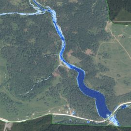 Das Bild zeigt eine Luftaufnahme einer Landschaft mit einem Fluss, der sich in einem geschwungenen Verlauf durch das Gebiet zieht. Der Fluss ist in kräftigem Blau dargestellt, während die umliegenden Flächen grün und braun gefärbt sind, was auf Wiesen, Felder und bewaldete Bereiche hinweist. Entlang des Flusses sind Straßen und vereinzelte Strukturen erkennbar, die auf eine ländliche Umgebung mit Infrastruktur schließen lassen.