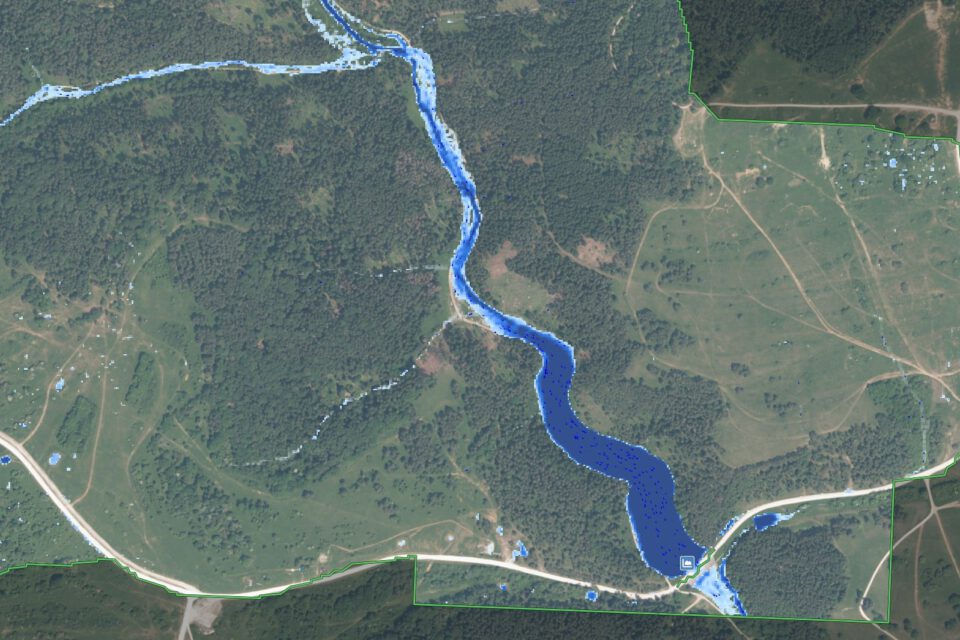 Das Bild zeigt eine Luftaufnahme einer Landschaft mit einem Fluss, der sich in einem geschwungenen Verlauf durch das Gebiet zieht. Der Fluss ist in kräftigem Blau dargestellt, während die umliegenden Flächen grün und braun gefärbt sind, was auf Wiesen, Felder und bewaldete Bereiche hinweist. Entlang des Flusses sind Straßen und vereinzelte Strukturen erkennbar, die auf eine ländliche Umgebung mit Infrastruktur schließen lassen.