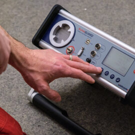 Das Bild zeigt eine Hand, die ein tragbares elektronisches Messgerät auf dem Boden bedient. Das Gerät verfügt über ein Display, mehrere Anschlüsse und Bedientasten. Die Szene wirkt wie eine technische Messung in einem Innenraum.