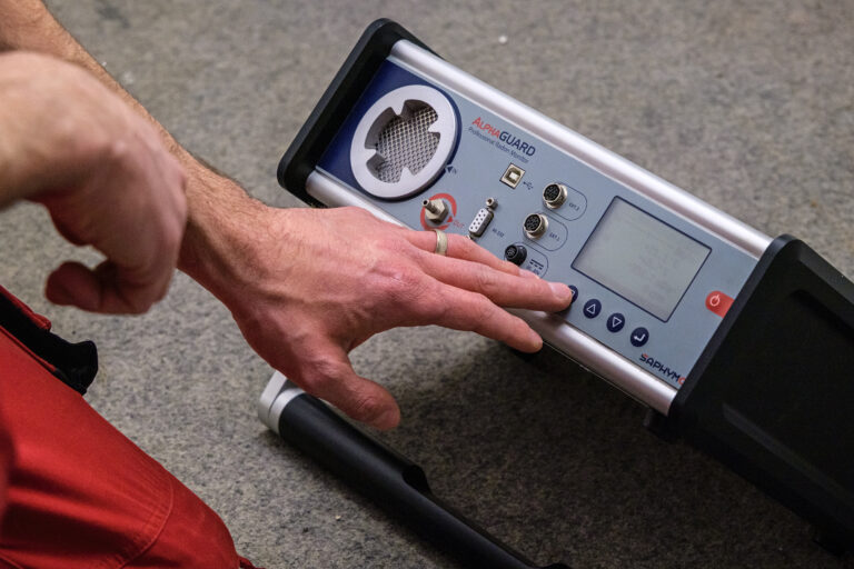 Das Bild zeigt eine Hand, die ein tragbares elektronisches Messgerät auf dem Boden bedient. Das Gerät verfügt über ein Display, mehrere Anschlüsse und Bedientasten. Die Szene wirkt wie eine technische Messung in einem Innenraum.