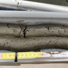 Das Bild zeigt eine Nahaufnahme eines Sedimentkerns aus grauem Schlamm oder Ton in einem zylindrischen Behälter. Unter dem Behälter liegt ein Maßband, das eine Länge des Kerns von etwa 45 bis 70 Zentimeter anzeigt.