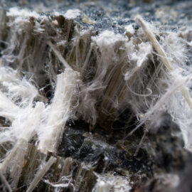 Das Bild zeigt eine extreme Nahaufnahme eines stark faserigen, grauen Materials mit zahlreichen feinen, weißen Fasern, die aus der Oberfläche herausragen. Die Struktur wirkt brüchig und unregelmäßig, mit klar erkennbaren Schichtungen im Inneren.
