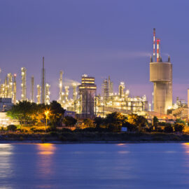 Das Bild zeigt eine große Industrieanlage bei Dämmerung oder Nacht, die hell beleuchtet ist. Mehrere hohe Türme und komplexe Rohrsysteme sind sichtbar, zusammen mit einem markanten Schornstein, der in den Himmel ragt. Im Vordergrund befindet sich ein ruhiger Wasserbereich, der die Lichter der Anlage leicht reflektiert, während der Hintergrund einen klaren, violett-blauen Himmel zeigt.