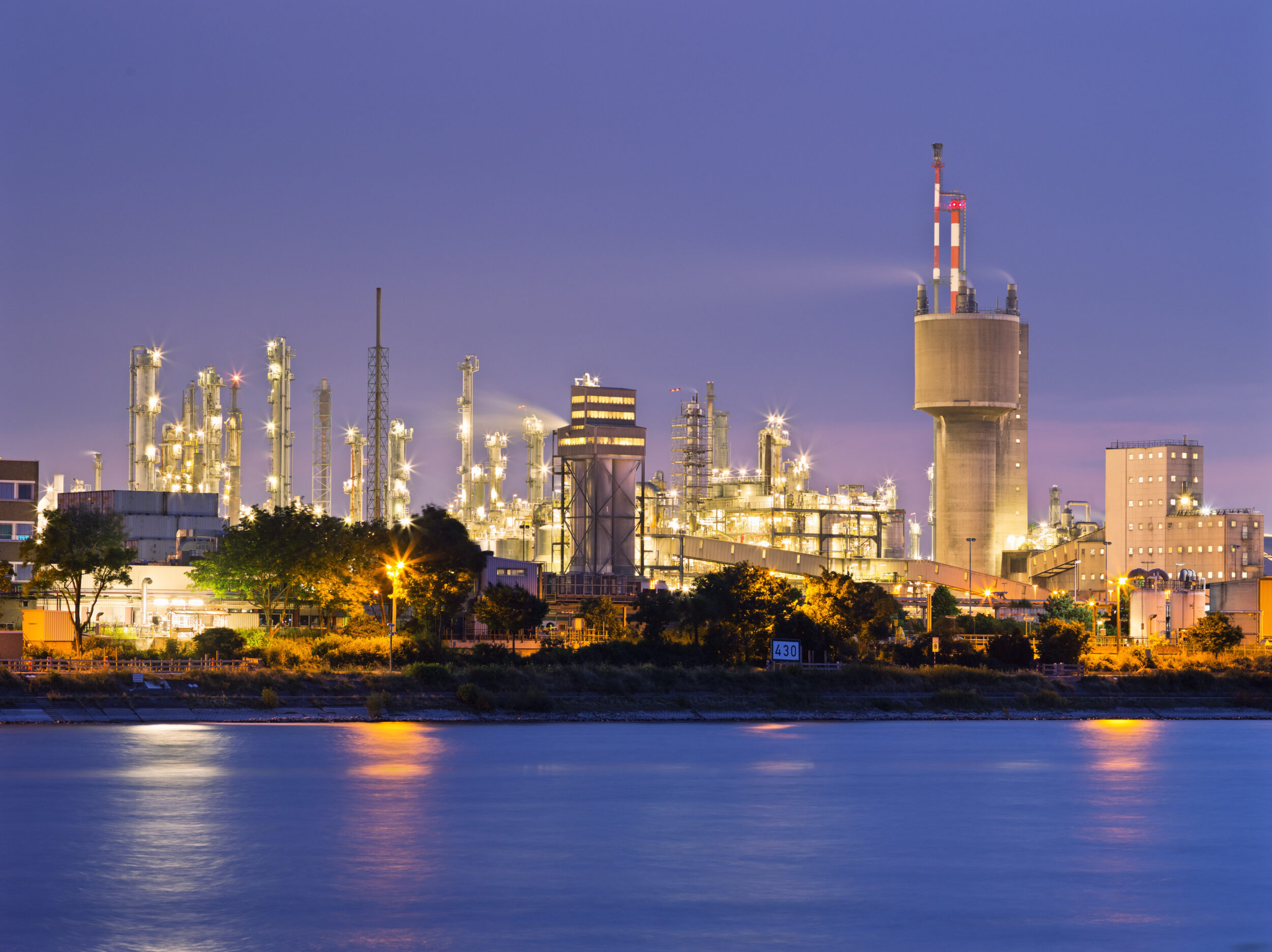 Das Bild zeigt eine große Industrieanlage bei Dämmerung oder Nacht, die hell beleuchtet ist. Mehrere hohe Türme und komplexe Rohrsysteme sind sichtbar, zusammen mit einem markanten Schornstein, der in den Himmel ragt. Im Vordergrund befindet sich ein ruhiger Wasserbereich, der die Lichter der Anlage leicht reflektiert, während der Hintergrund einen klaren, violett-blauen Himmel zeigt.