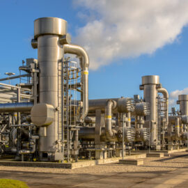 Das Bild zeigt eine große industrielle Anlage im Freien mit mehreren massiven Metallkonstruktionen und Rohrsystemen. Die Anlage besteht aus hohen, zylindrischen Türmen und zahlreichen miteinander verbundenen Leitungen, die auf eine Gas- oder Chemieverarbeitungsstation hinweisen. Der Boden ist gepflastert, und im Hintergrund ist ein klarer Himmel mit einigen Wolken zu sehen, was die Szene hell und technisch geprägt erscheinen lässt.