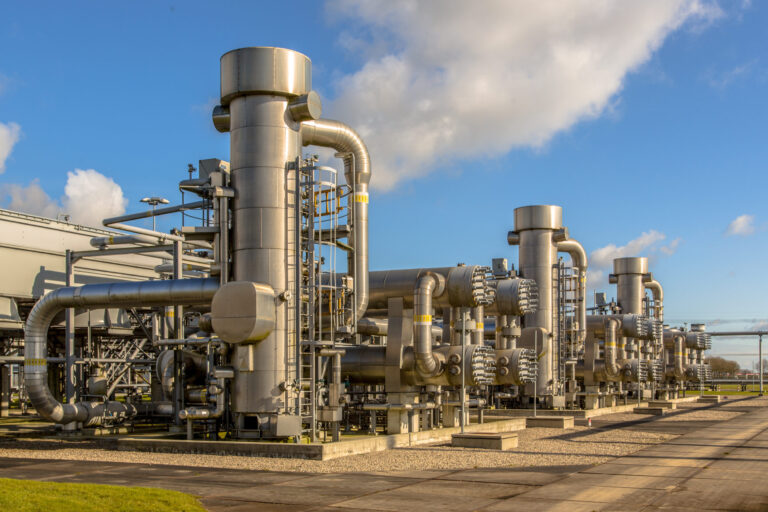 Das Bild zeigt eine große industrielle Anlage im Freien mit mehreren massiven Metallkonstruktionen und Rohrsystemen. Die Anlage besteht aus hohen, zylindrischen Türmen und zahlreichen miteinander verbundenen Leitungen, die auf eine Gas- oder Chemieverarbeitungsstation hinweisen. Der Boden ist gepflastert, und im Hintergrund ist ein klarer Himmel mit einigen Wolken zu sehen, was die Szene hell und technisch geprägt erscheinen lässt.