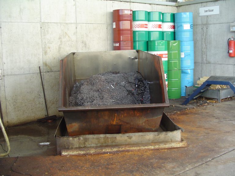 Das Bild zeigt einen Innenbereich mit einer großen, offenen Metallwanne im Vordergrund, die mit metallischen Spänen und Restmaterial gefüllt ist. Dahinter stehen mehrere farbige Metallfässer in Rot, Grün und Blau, die vermutlich für die Lagerung von Flüssigkeiten oder Ölen verwendet werden. Die Umgebung wirkt wie ein Industrie- oder Werkstattbereich mit Betonwänden, einem Feuerlöscher an der Wand und einer leicht erhöhten Plattform auf der rechten Seite.
