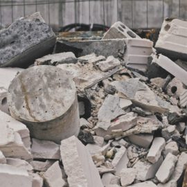 Das Bild zeigt einen Container, der mit einer großen Menge an Bauschutt gefüllt ist. Darin befinden sich verschiedene Materialien wie zerbrochene Betonstücke, Ziegelsteine und ein zylindrisches Betonteil. Die Bruchstücke sind überwiegend hellgrau und weiß, mit rauen und unregelmäßigen Kanten, und der Container besteht aus rostigem Metall.