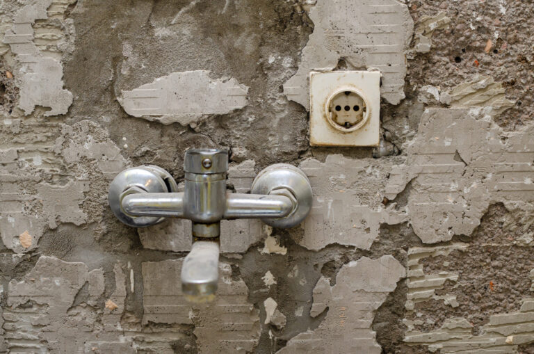 Das Bild zeigt eine raue, graue Wand mit teilweise abgeplatztem Putz, wodurch die darunterliegende Struktur sichtbar wird. In der Mitte ist eine verchromte Armatur montiert, die aus zwei seitlichen Anschlüssen und einem zentralen Hebel besteht. Rechts oberhalb der Armatur befindet sich eine alte, vergilbte Steckdose, die in die beschädigte Wand eingelassen ist.