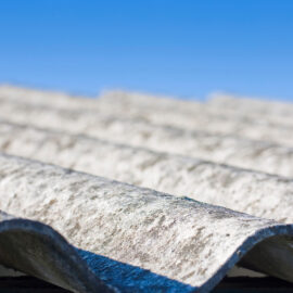 Das Bild zeigt ein Dach mit wellenförmigen Platten aus hellem, verwittertem Material, das deutliche Alterungsspuren aufweist. Die Oberfläche wirkt rau und unregelmäßig, mit Verfärbungen und kleinen Ablagerungen. Im Hintergrund ist ein klarer blauer Himmel zu sehen, der die Struktur des Daches deutlich hervorhebt.