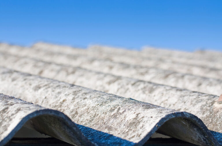 Das Bild zeigt ein Dach mit wellenförmigen Platten aus hellem, verwittertem Material, das deutliche Alterungsspuren aufweist. Die Oberfläche wirkt rau und unregelmäßig, mit Verfärbungen und kleinen Ablagerungen. Im Hintergrund ist ein klarer blauer Himmel zu sehen, der die Struktur des Daches deutlich hervorhebt.