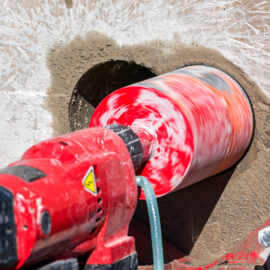Das Bild zeigt eine rot-weiße Kernbohrmaschine, die ein rundes Loch in eine Betonwand bohrt. Der Bohrkopf ist groß und zylindrisch, mit einer auffälligen rot-weißen Musterung, und er dreht sich sichtbar schnell, wodurch die Farben verschwimmen. Rund um die Bohrstelle ist feiner Betonstaub zu sehen, und ein grüner Schlauch führt Wasser zur Kühlung des Bohrvorgangs.