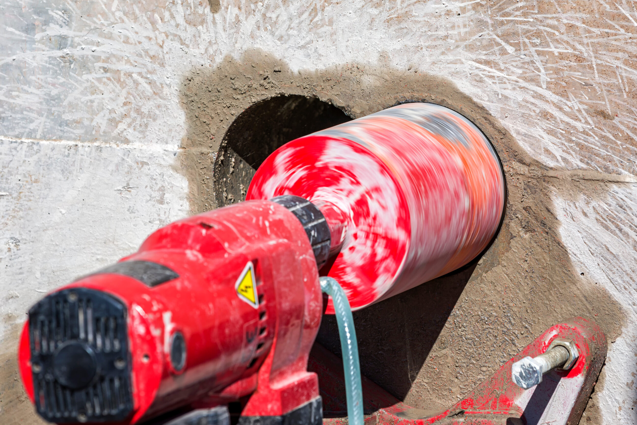 Das Bild zeigt eine rot-weiße Kernbohrmaschine, die ein rundes Loch in eine Betonwand bohrt. Der Bohrkopf ist groß und zylindrisch, mit einer auffälligen rot-weißen Musterung, und er dreht sich sichtbar schnell, wodurch die Farben verschwimmen. Rund um die Bohrstelle ist feiner Betonstaub zu sehen, und ein grüner Schlauch führt Wasser zur Kühlung des Bohrvorgangs.