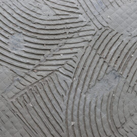 Das Bild zeigt eine graue Oberfläche, die mit einem Muster aus tiefen Rillen und diagonalen Linien versehen ist. Die Rillen verlaufen in geschwungenen Bögen und überkreuzen sich, wodurch ein strukturiertes, fast dekoratives Erscheinungsbild entsteht. Zwischen den Rillen sind kleine quadratische Vertiefungen sichtbar, die auf eine vorbereitete Fläche für Fliesen oder Bodenbelag hindeuten.