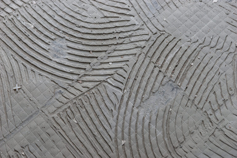 Das Bild zeigt eine graue Oberfläche, die mit einem Muster aus tiefen Rillen und diagonalen Linien versehen ist. Die Rillen verlaufen in geschwungenen Bögen und überkreuzen sich, wodurch ein strukturiertes, fast dekoratives Erscheinungsbild entsteht. Zwischen den Rillen sind kleine quadratische Vertiefungen sichtbar, die auf eine vorbereitete Fläche für Fliesen oder Bodenbelag hindeuten.