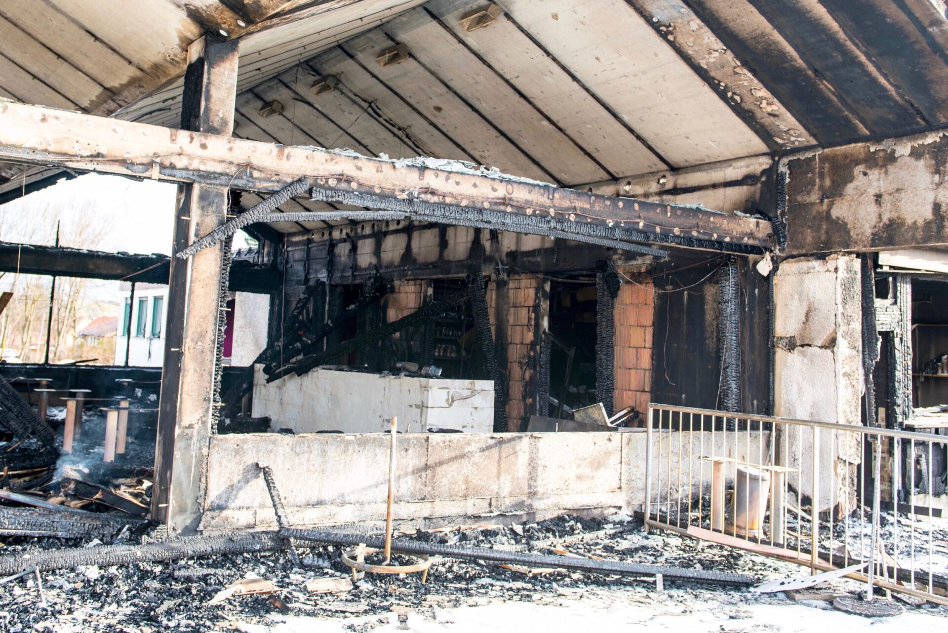 Das Bild zeigt den stark beschädigten Innenbereich eines Gebäudes nach einem Brand. Die Decke und die tragenden Balken sind stark verrußt und teilweise verkohlt, während die Wände deutliche Brandspuren und freiliegende Mauersteine zeigen. Auf dem Boden liegen verbrannte Trümmer und verkohlte Reste von Möbeln, und im Hintergrund sind dunkle, ausgebrannte Räume erkennbar.