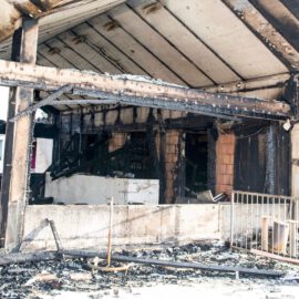 Das Bild zeigt den stark beschädigten Innenbereich eines Gebäudes nach einem Brand. Die Decke und die tragenden Balken sind stark verrußt und teilweise verkohlt, während die Wände deutliche Brandspuren und freiliegende Mauersteine zeigen. Auf dem Boden liegen verbrannte Trümmer und verkohlte Reste von Möbeln, und im Hintergrund sind dunkle, ausgebrannte Räume erkennbar.