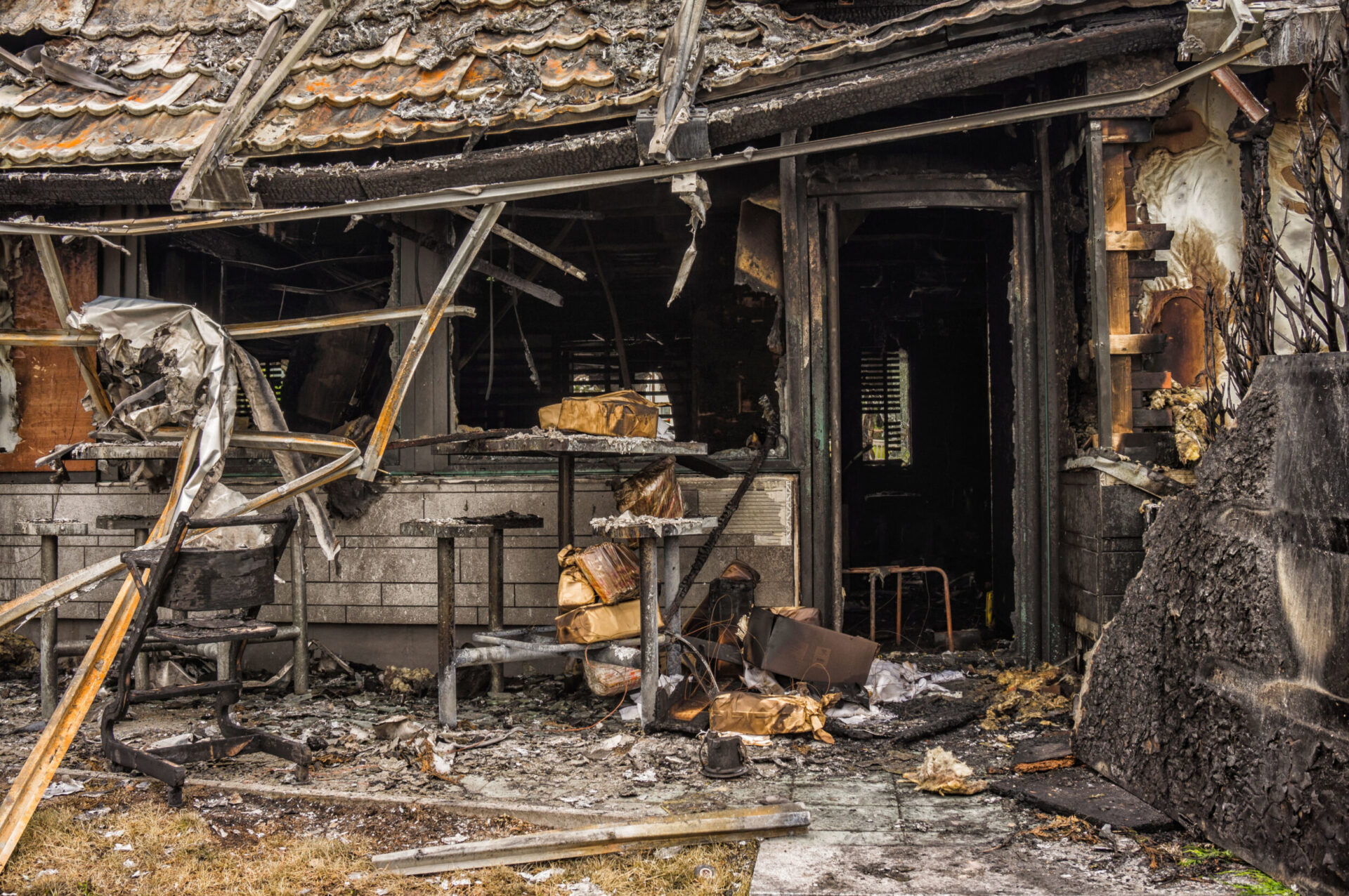Das Bild zeigt die stark beschädigte Fassade eines Gebäudes nach einem Brand. Die Wände sind verrußt und teilweise eingestürzt, mit verbrannten Holz- und Metallteilen, die chaotisch hervorstehen. Im Vordergrund liegen verkohlte Möbelreste und Trümmer, während der Innenraum dunkel und ausgebrannt wirkt, mit sichtbaren Spuren von Feuer und Rauch.