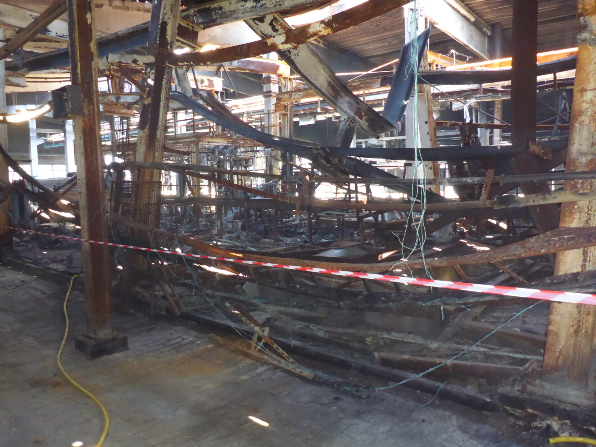 Das Bild zeigt den Innenbereich einer stark beschädigten Industriehalle nach einem Brand. Die Metallkonstruktionen sind stark verbogen und teilweise eingestürzt, viele Flächen sind verbrannt und mit Ruß bedeckt. Im Vordergrund ist ein rot-weißes Absperrband gespannt, das den Bereich sichert, während im Hintergrund verkohlte Reste von Maschinen und Leitungen sichtbar sind.