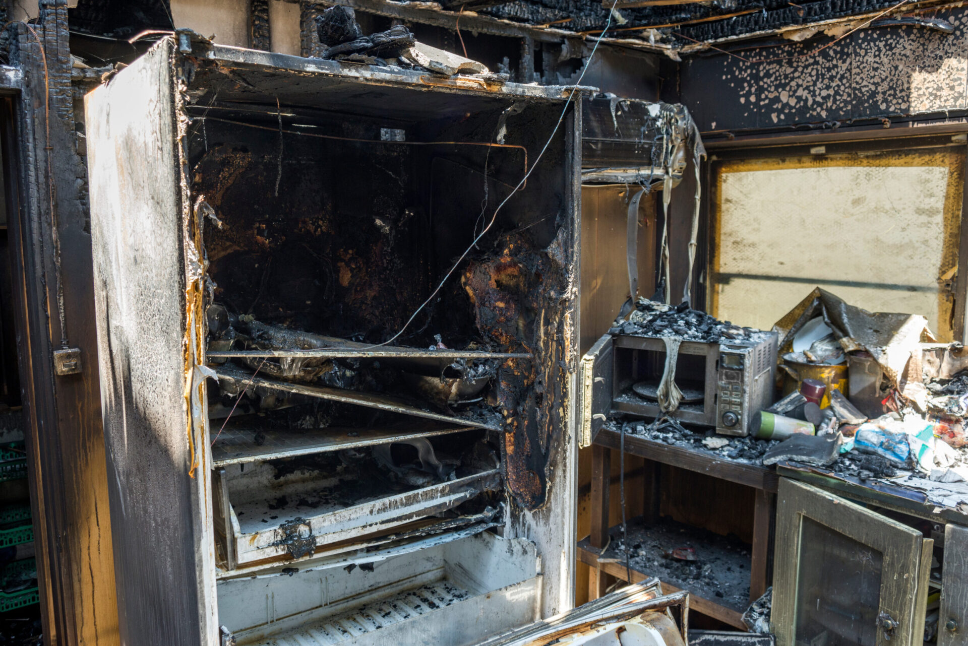 Das Bild zeigt eine stark beschädigte Küche nach einem Brand. Ein großer Kühlschrank ist vollständig verbrannt und verkohlt, ebenso sind die Wände und Decke stark verrußt. Auf der Arbeitsfläche liegen verbrannte Reste von Küchengeräten und Materialien, die Umgebung ist mit Asche und Trümmern bedeckt.
