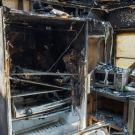 Das Bild zeigt eine stark beschädigte Küche nach einem Brand. Ein großer Kühlschrank ist vollständig verbrannt und verkohlt, ebenso sind die Wände und Decke stark verrußt. Auf der Arbeitsfläche liegen verbrannte Reste von Küchengeräten und Materialien, die Umgebung ist mit Asche und Trümmern bedeckt.