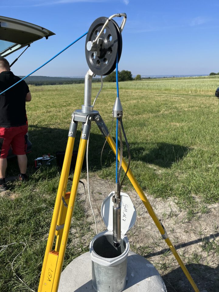 Das Bild zeigt ein Messgerät, das auf einem gelben Stativ über einem Brunnenrohr installiert ist. An dem Gerät ist ein Kabel befestigt, das in das geöffnete Rohr führt, vermutlich für eine Inspektion oder Messung. Im Hintergrund ist eine weitläufige Wiese unter blauem Himmel zu sehen, und am linken Bildrand steht eine Person neben einem Fahrzeug.
