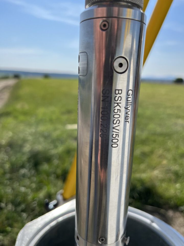 Das Bild zeigt eine Nahaufnahme eines metallischen Messgeräts, das senkrecht in ein Brunnenrohr eingesetzt ist. Auf der glänzenden Oberfläche des Geräts sind feine Schrauben und technische Details erkennbar, die auf eine präzise Verarbeitung hinweisen. Im Hintergrund ist unscharf eine grüne Wiese unter blauem Himmel zu sehen, was auf eine Messung im Freien hindeutet.