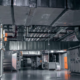 Das Bild zeigt einen großen Technikraum mit umfangreicher Lüftungs- und Klimaanlagentechnik. An der Decke verlaufen zahlreiche rechteckige und runde Lüftungskanäle aus Metall, die teilweise mit Isolierung versehen sind. Im Vordergrund und an den Seiten sind Rohrleitungen, Steuerkästen und orangefarbene Bedienelemente sichtbar, während der Boden aus glattem grauem Material besteht und die gesamte Szene einen industriellen Charakter hat.