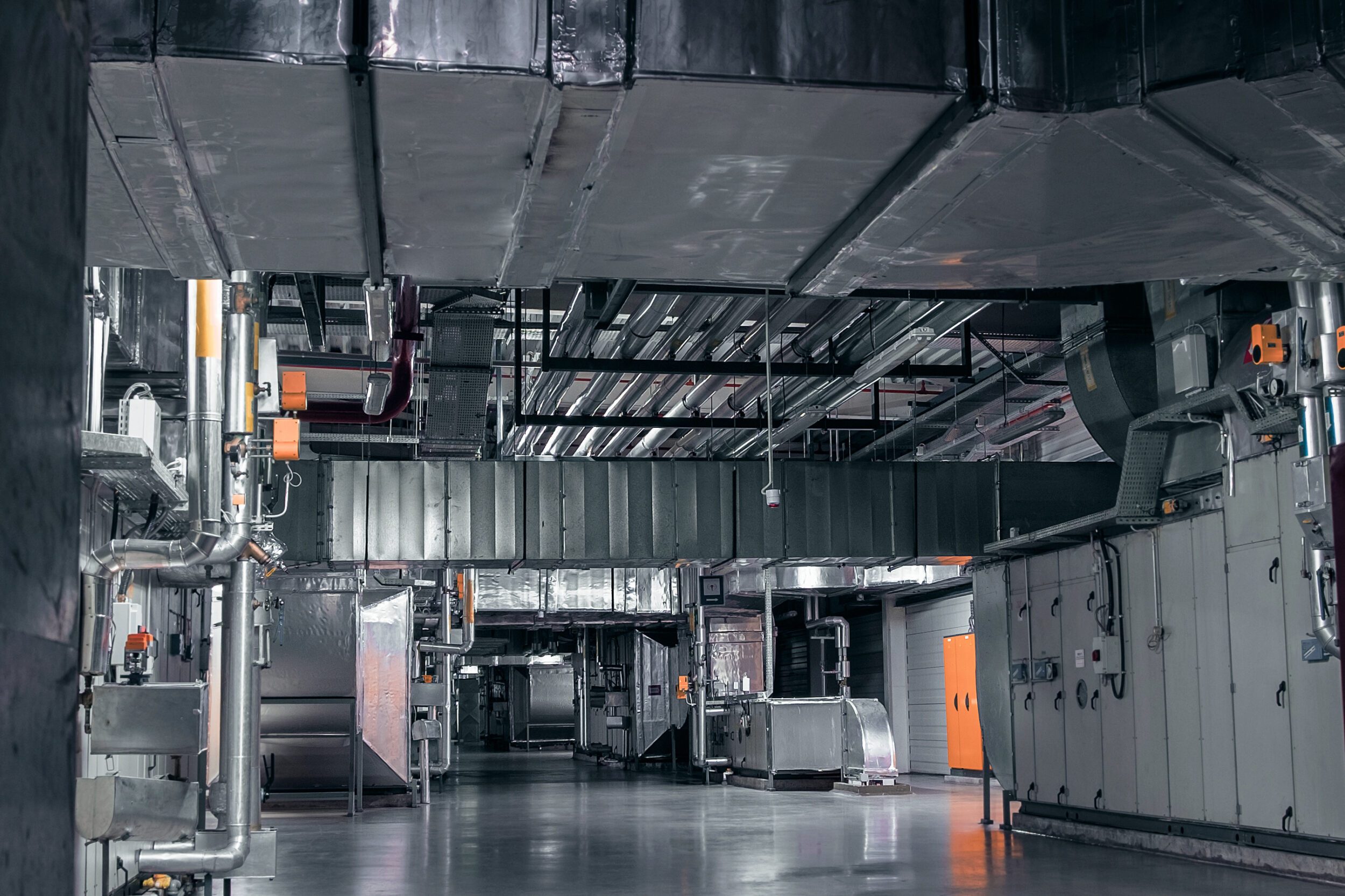 Das Bild zeigt einen großen Technikraum mit umfangreicher Lüftungs- und Klimaanlagentechnik. An der Decke verlaufen zahlreiche rechteckige und runde Lüftungskanäle aus Metall, die teilweise mit Isolierung versehen sind. Im Vordergrund und an den Seiten sind Rohrleitungen, Steuerkästen und orangefarbene Bedienelemente sichtbar, während der Boden aus glattem grauem Material besteht und die gesamte Szene einen industriellen Charakter hat.