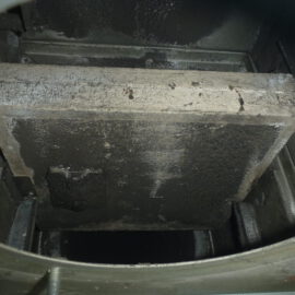 Das Bild zeigt das Innere einer Brandschutzklappe in einem Lüftungssystem. In der Mitte befindet sich eine große rechteckige Klappe aus Metall, die stark verschmutzt und mit dunklen Ablagerungen bedeckt ist. Die umliegenden Flächen sind ebenfalls dunkel verfärbt, was auf Ruß oder Staubablagerungen hinweist, und die Mechanik der Klappe ist teilweise sichtbar.