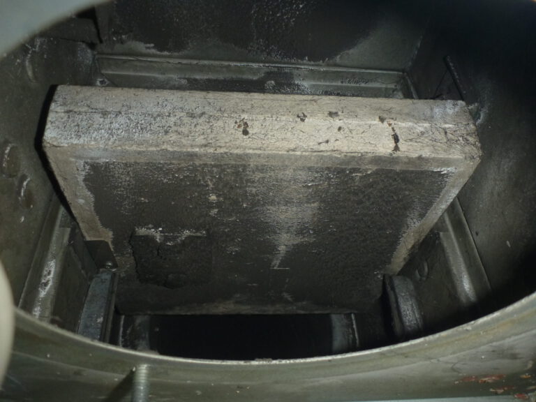 Das Bild zeigt das Innere einer Brandschutzklappe in einem Lüftungssystem. In der Mitte befindet sich eine große rechteckige Klappe aus Metall, die stark verschmutzt und mit dunklen Ablagerungen bedeckt ist. Die umliegenden Flächen sind ebenfalls dunkel verfärbt, was auf Ruß oder Staubablagerungen hinweist, und die Mechanik der Klappe ist teilweise sichtbar.