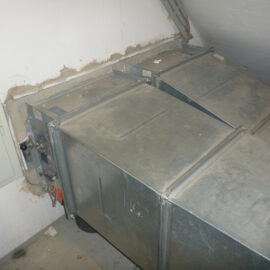 Das Bild zeigt eine große metallische Lüftungsanlage, die in einem Technikraum installiert ist. Die Anlage besteht aus mehreren rechteckigen Kanälen mit glatten Oberflächen, die an einer Wand befestigt sind. An der linken Seite sind Kabel und Anschlüsse sichtbar, während die Wand rund um die Öffnung Spuren von Abdichtmaterial aufweist.